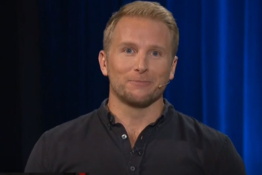 A man with blond hair in front of a curtain, wears a microphone.