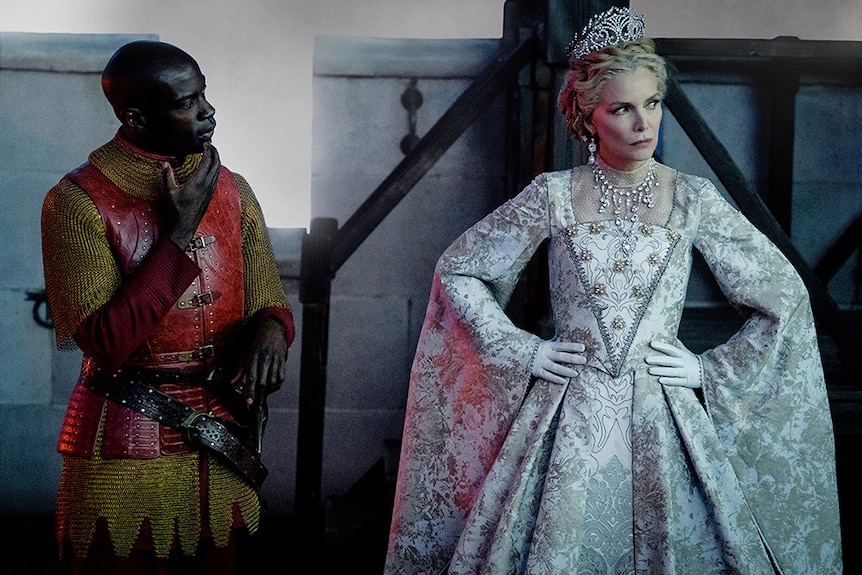 David Gyasi in red and gold armour stands looking to Michelle Pfeiffer with hands on hips in crown and ornate jewelled gown.