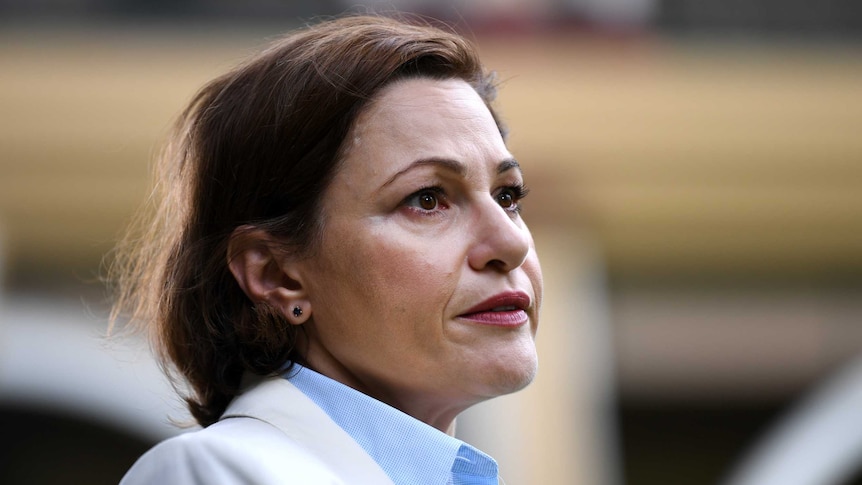 Jackie Trad looks to the right. Her brown hair is down, she wears a beige blazer and pale blue shirt and has red lipstick on.