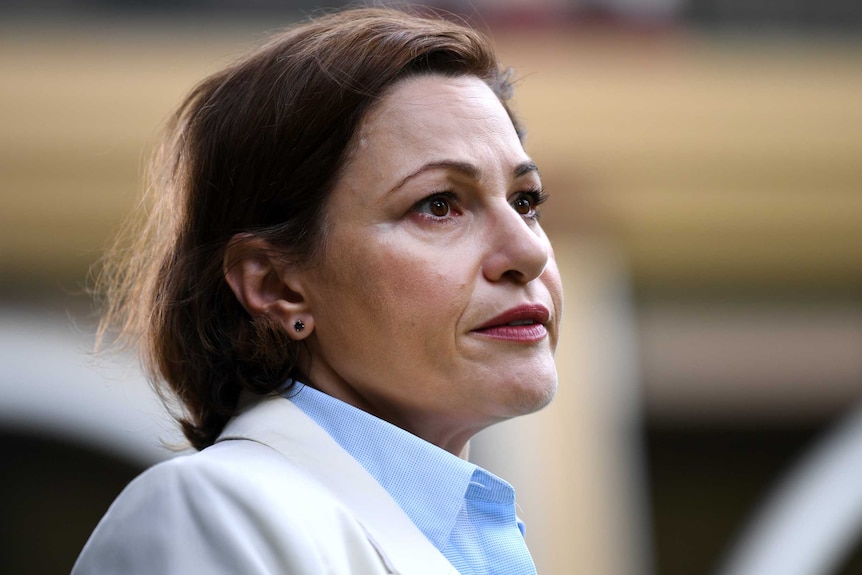 Jackie Trad looks to the right. Her brown hair is down, she wears a beige blazer and pale blue shirt and has red lipstick on.