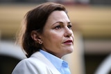 Jackie Trad looks to the right. Her brown hair is down, she wears a beige blazer and pale blue shirt and has red lipstick on.