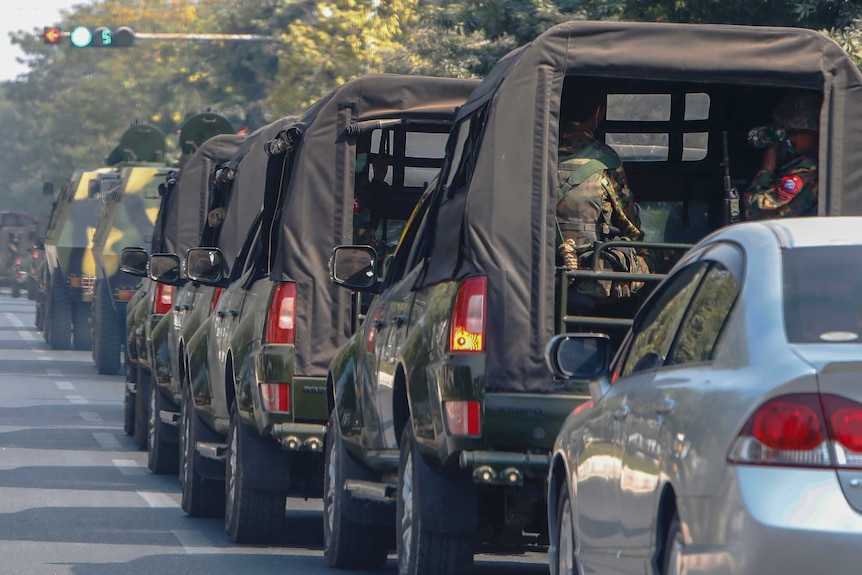 A convoy of eight army vehicles rolls down a long street in daylight, with armed soldiers in the back of some 4WDs