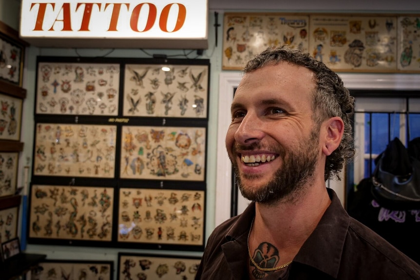 A man smiles in front of a wall of tattoo designs, with a lit sign saying 'tattoo' in the background. 