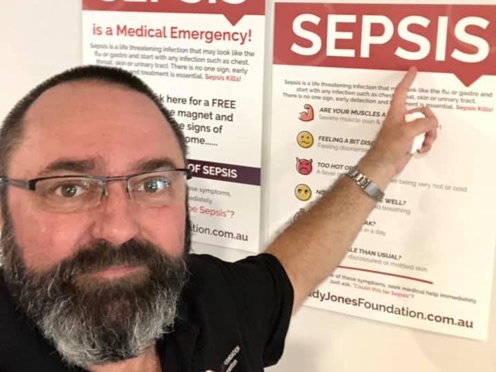 A man with a dark beard and glasses pointing to a Sepsis sign on the wall