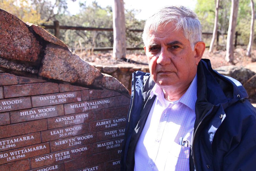 A man with white hear, wearing a checked shirt and blue jacket, stands next to a wall with names inscribed on it.