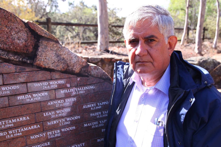 A man with white hear, wearing a checked shirt and blue jacket, stands next to a wall with names inscribed on it.