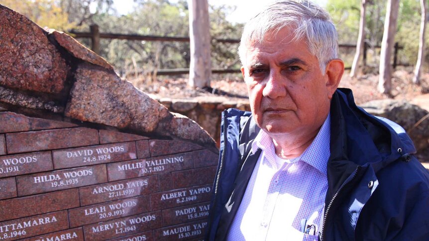 A man with white hear, wearing a checked shirt and blue jacket, stands next to a wall with names inscribed on it.