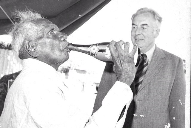An older Aboriginal man takes a sip from a champagne bottle as a white man looks on smiling.