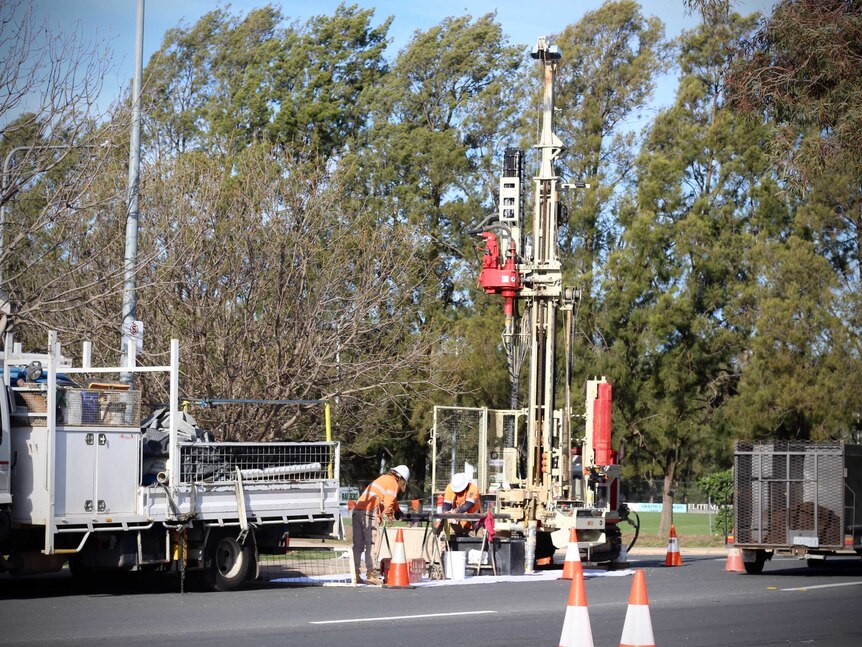 Workers in high vis operate a large drill near a truck on the side of the road.
