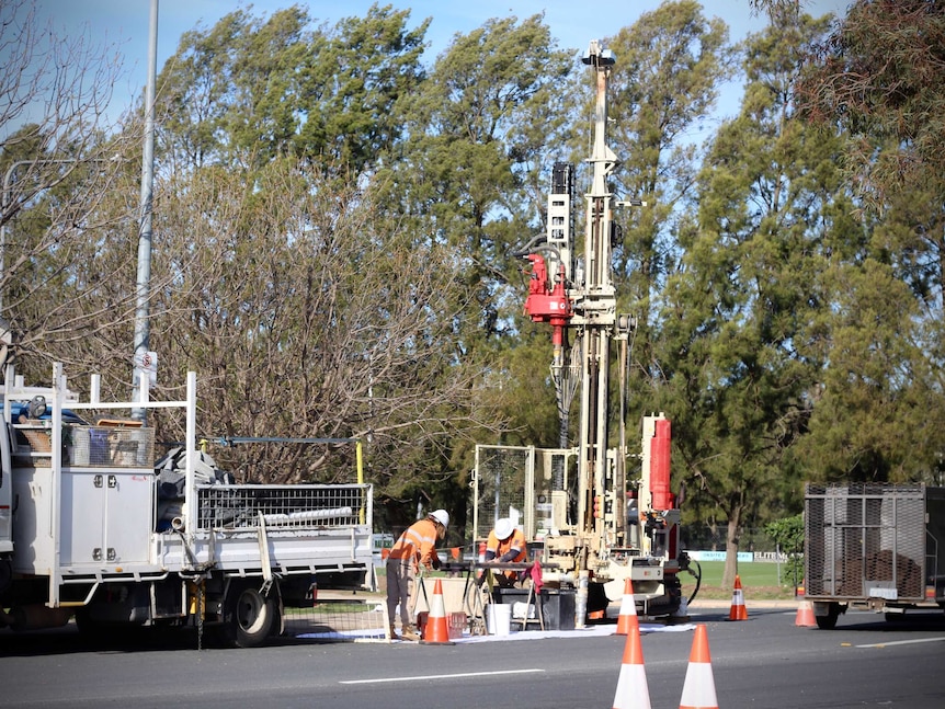 Workers in high vis operate a large drill near a truck on the side of the road.