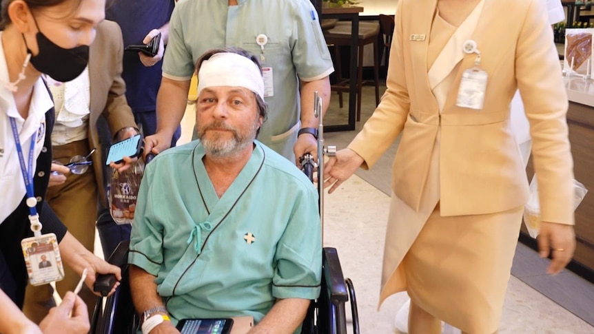 A man with his head bandaged and in a wheelchair being whisked away and surrounded by people
