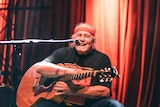 Kev Carmody wearing a red headband, holding a guitar, laughing