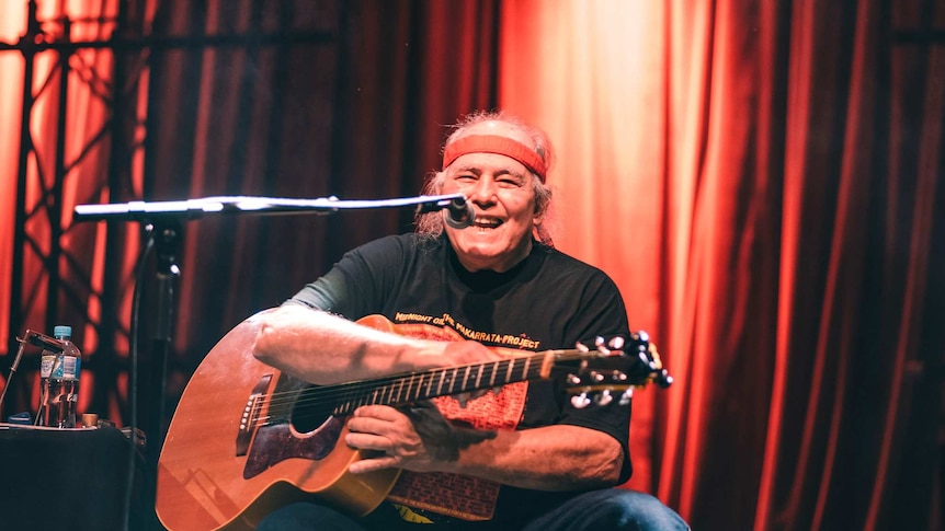 Kev Carmody wearing a red headband, holding a guitar, laughing