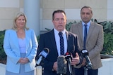 A blonde woman in sky blue suit and a man in beige suit, stand behind a man in a dark suit who speaks at a press conference.