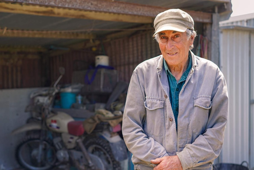 An older man in a grey cap smiles at the camera, a motorbike and shed behind him.