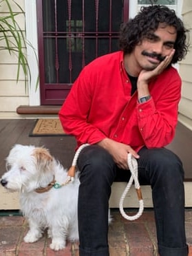 A young man sits on the front step of a house, contentedly resting his face on his hand, a small fluffy dog by his side.
