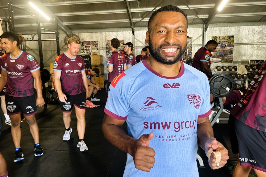 A man from Papua New Guinea wearing a light blue training shirt smiling as his teammates work out in the gym