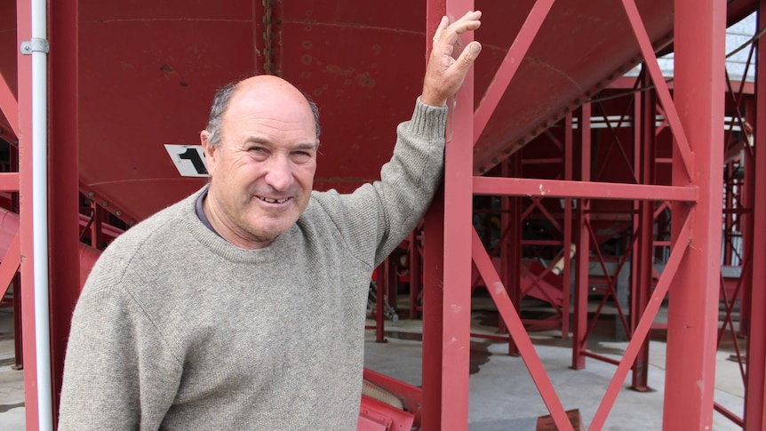 Man in grey jumper stands in foreground, leaning against  red grain silo