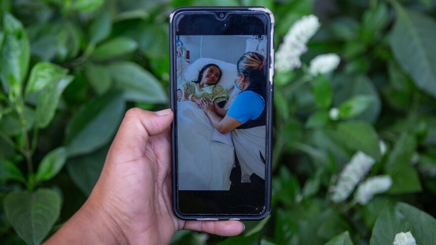 A person's hand holding a phone in front of green leaves, which displays a photo of a girl in a hospital bed 