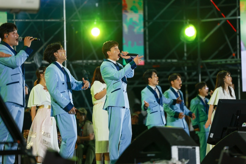 Six men in matching blue suits sing enthusiastically into microphones on stage