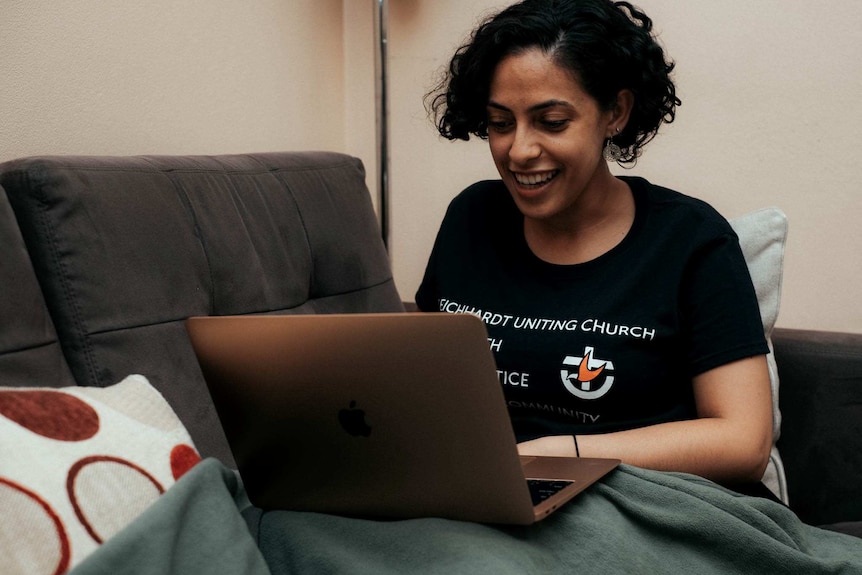Christina Mikhael on couch, wearing Uniting Church t-shirt and looking at laptop.