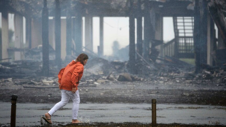 Woman in orange jacket walks in front of burned house