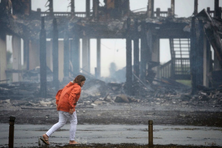 Woman in orange jacket walks in front of burned house