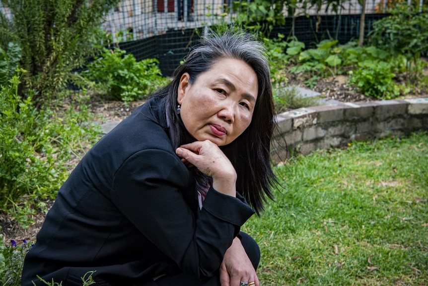 A woman with long dark hair sits in a garden with her hand on her chin.