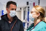 A man with a surgical face mask on speaking to a man and a woman who are wearing hospital scrubs 
