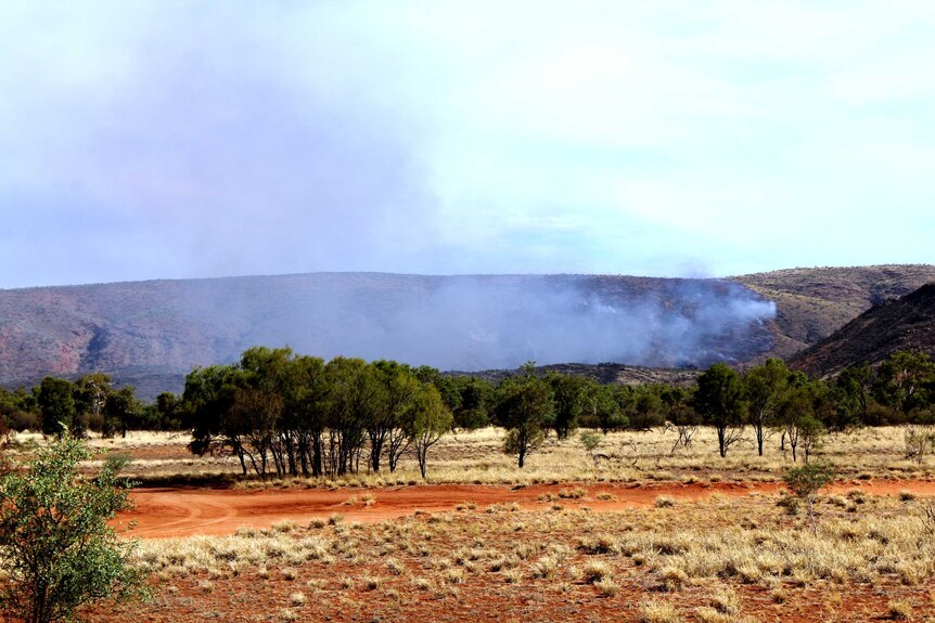 A long, wide shot of a mountain range and smoke from a dwindling fire. Bush and red dirt in the foreground.