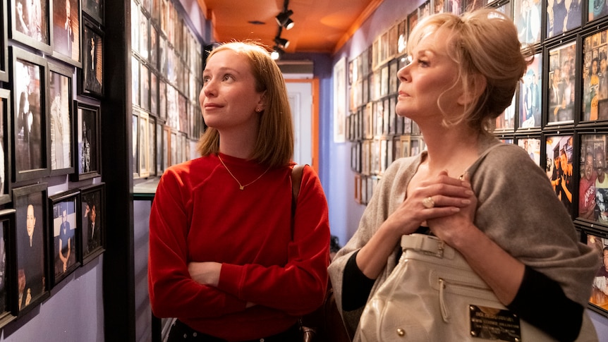 A 25 year old woman with red hair stands next to a woman in her late 60s with blonde hair, they are looking at photos in a hall