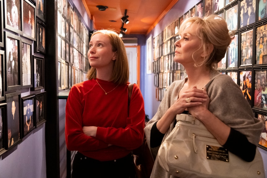 A 25 year old woman with red hair stands next to a woman in her late 60s with blonde hair, they are looking at photos in a hall