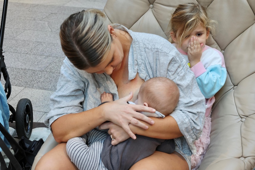 A woman breastfeeding her child, with a young girl sitting behind her.