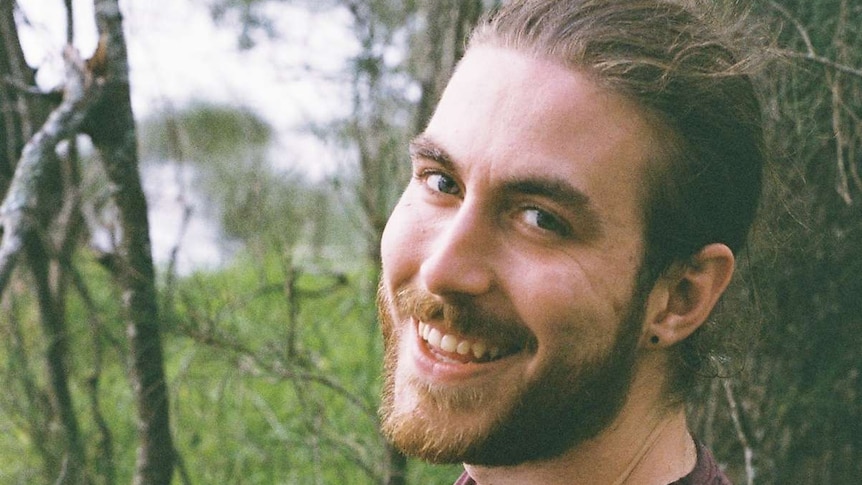 Jack Heskett, a young person with a beard, sits outside in the bush and smiles towards the camera