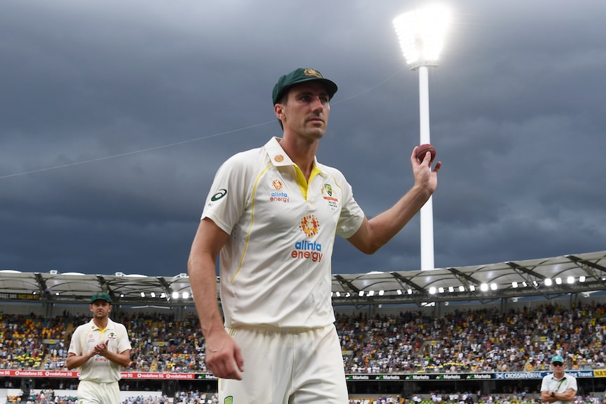 Australian bowler Pat Cummins holds a cricket ball in the air in a stadium. the sky is dark with storm clouds