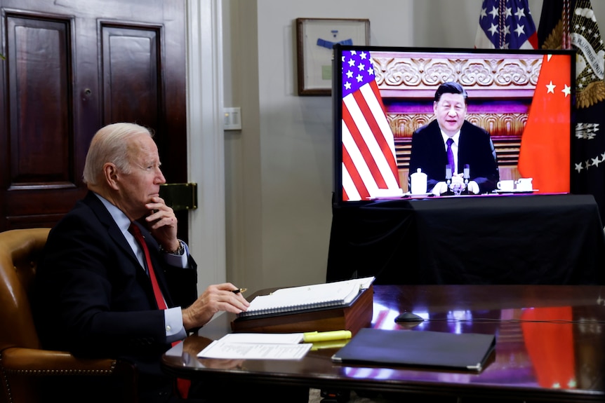 Joe Biden sits at a desk while Xi Jinping can be seen on a TV screen.