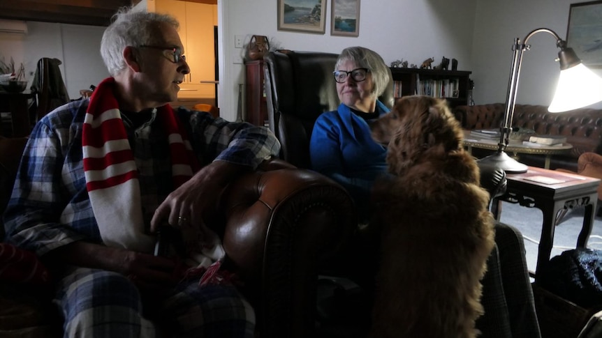 A woman and a man with a dog in a living room with one small lamp.