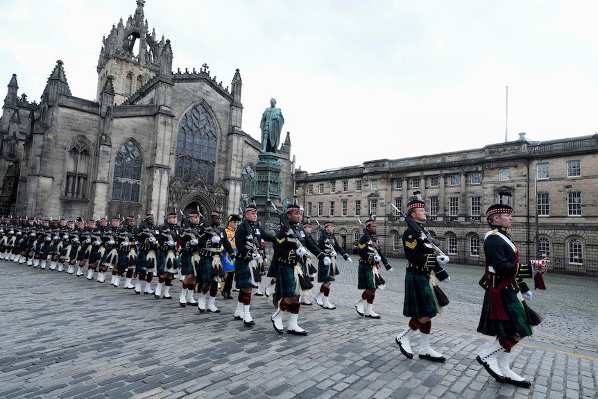 Guards in green tartan kilts and white boots march in formation outside a large, old Cathedral