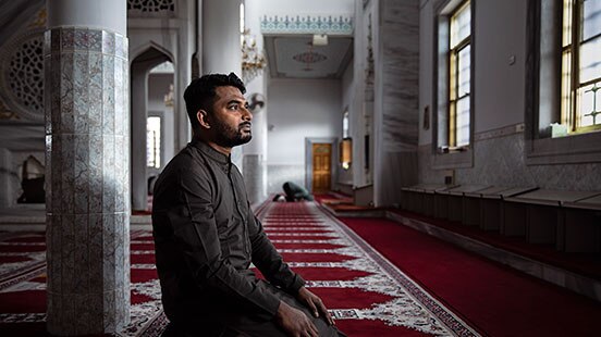 Muslim man Khaled Shaikh kneeling on carpet in mosque.