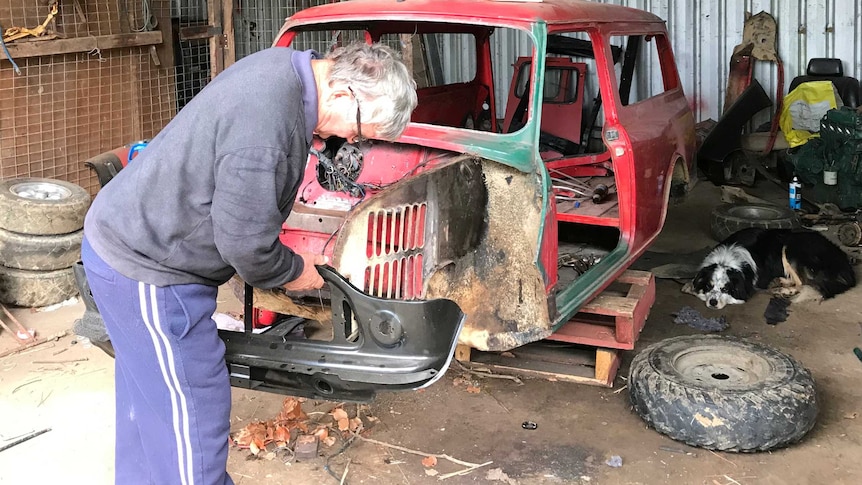 An elderly man works on an old car in a farm shed