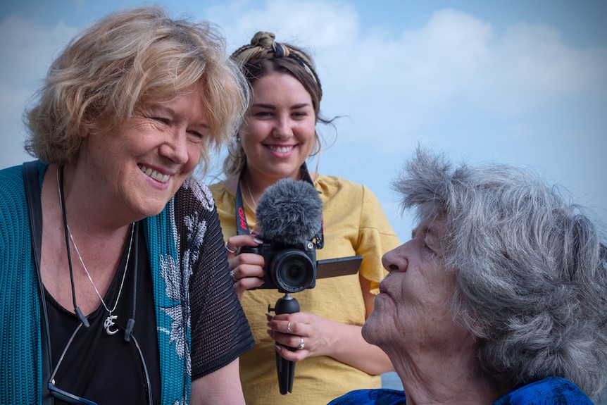 Three women of different generations. The two older women are smiling at each other and the youngest is filming the moment.