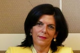 Julia Banks, in yellow, sits on a chair with her legs crossed and arms folded in her lap, with a neutral expression on her face.