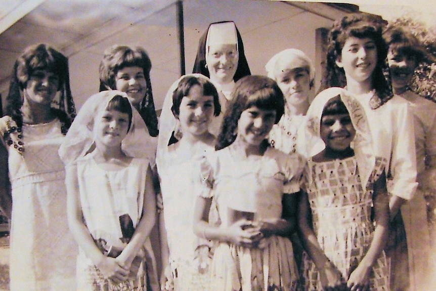 A group of children in dresses stand with a nun in full robes and headwear.