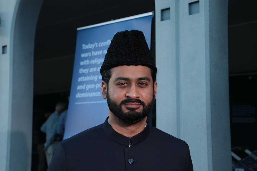 A man with a black beard wearing a black hat standing infront of a mosque