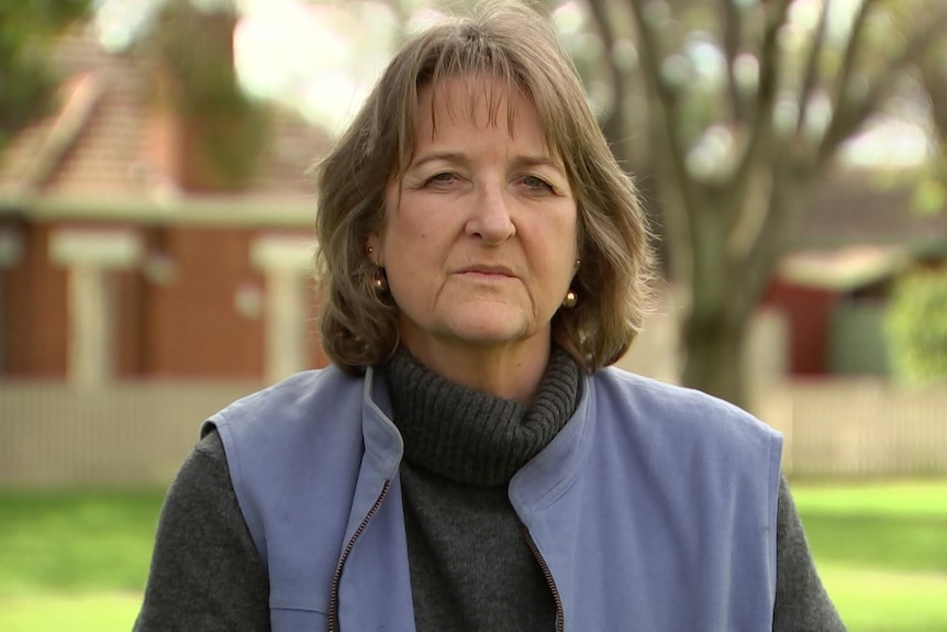 A woman with a grey turtleneck jumper and a blue fleece vest stands in a park, with houses in the background