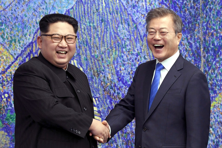 Two smiling men in formal wear shake hands as they pose for a photograph in front of a blue background.