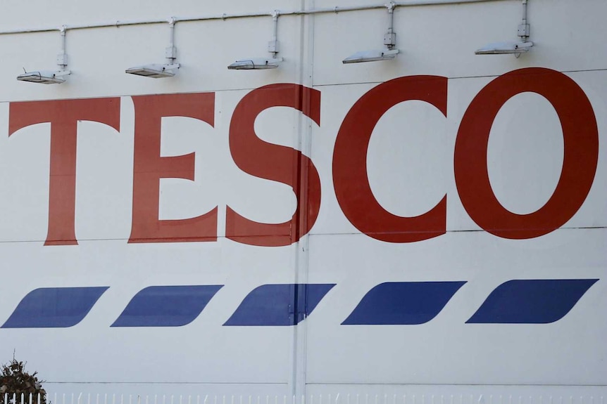 A man walks past a sign saying "Tesco" and "Helping you spend less everyday".