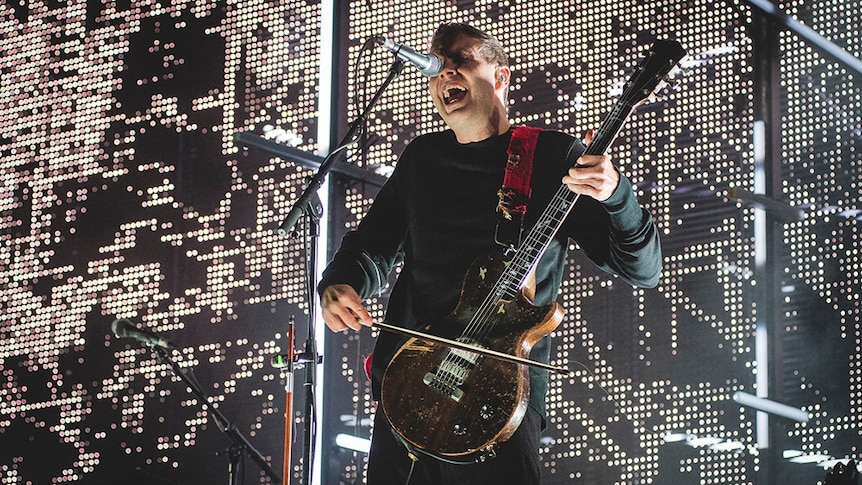 Jonsi of Sigur Ros bows a guitar and sings into a microphone on stage in front of a sheet of LED lights