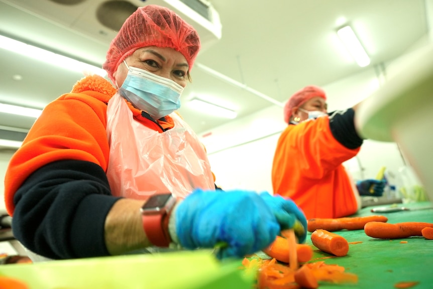 A woman wearing a mask, gloves and plastic apron sorts carrots.