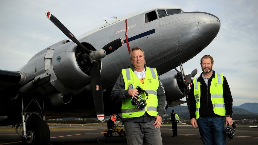 Two older men in high-vis stand holding aviation headphones in front of a DC-3 aircraft.